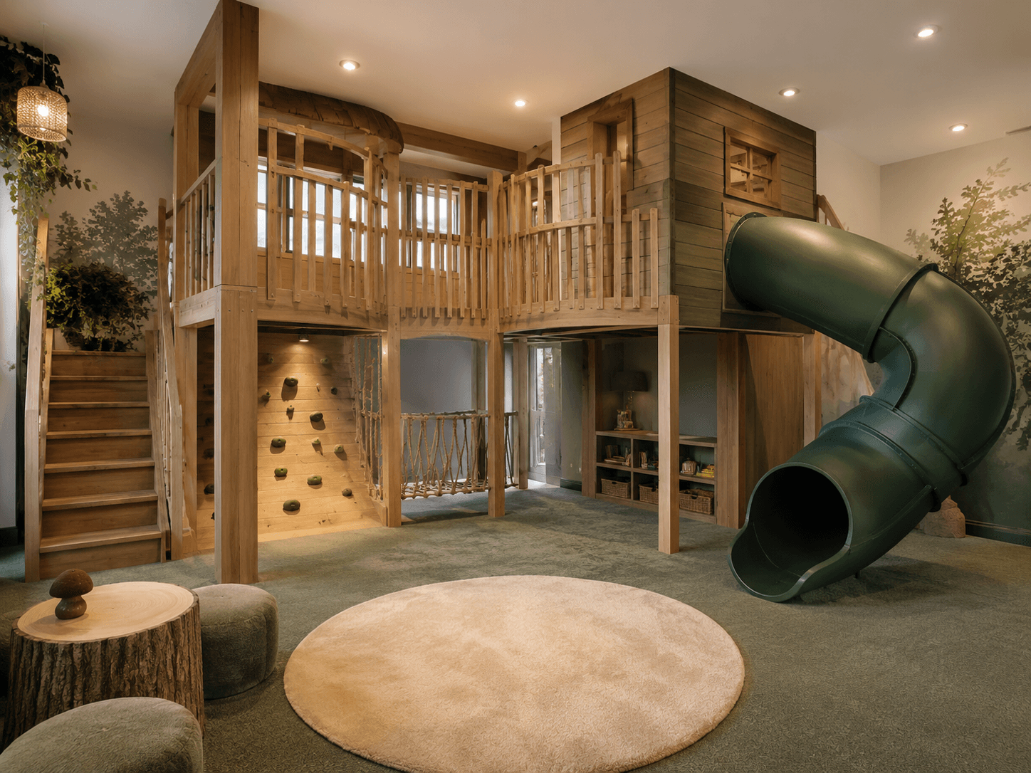 Two-story treehouse playroom with a green tube slide, climbing wall, rope bridge, and log stools on forest-green carpet